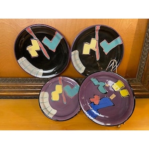 May include: A set of four ceramic plates featuring abstract geometric shapes in yellow, teal, blue, and pink. Two plates have a glossy black base, while the other two have a purple base. Each plate displays a grid-like pattern in white and blue.