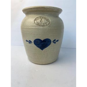May include: A beige stoneware crock featuring a blue painted heart design on the front. The upper portion of the vessel displays an embossed circular stamp with the text PINEWOOD VALLEY and a small tree icon centered within the lettering.