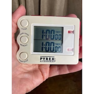 May include: A rectangular off-white digital kitchen timer with a dual display showing 1:00:00 for both Timer 1 and Timer 2. The device features buttons labeled Clear, 12HR/24HR, Memory, Start/Stop, T12, and Set, with the Pyrex Accessories logo printed below.