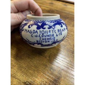 May include: A small, round, white ceramic jar with blue decorative patterns and text. The text reads "MAGDA TOILET CREAM C.U. COUNTIE & CO. CHEMISTS BOSTON U.S.A." The jar is held by a person.