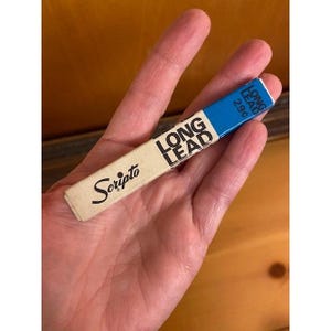 May include: A rectangular Scripto brand lead refill container featuring a cream-colored base and a blue top section. The text "LONG LEAD" is printed in black on the cream portion, while the blue section displays "LONG LEAD" and "29¢".