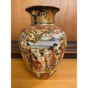 May include: A ceramic vase featuring a white base with intricate hand-painted scenes of figures in traditional attire near a body of water. The neck is decorated with dark blue and gold patterns, while the body displays colorful floral motifs.