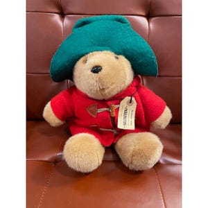 May include: A Paddington Bear plush toy wearing a green hat and a red duffle coat. The bear has a tag that reads "Paddington". The plush bear is sitting on a brown surface.