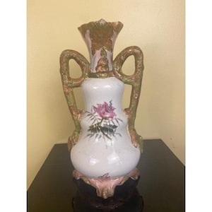 May include: A white ceramic vase with a pink and purple floral design. The vase has two handles and a flared top with gold and green accents. The base is pink and ornate.