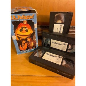 May include: A cardboard box set for H.R. Pufnstuf featuring a colorful illustration of the characters on the front, accompanied by three black VHS tapes labeled Volume 1, Volume 2, and Volume 3, each with white labels and black text.