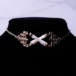 Puede incluir: Un collar gargantilla dorado con detalles de filigrana ornamentados y una delicada cadena. El punto focal presenta dos huesos blancos cruzados. El collar se exhibe en un maniqu&iacute; negro.