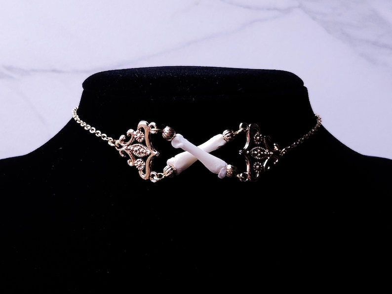 Puede incluir: Un collar gargantilla dorado con detalles de filigrana ornamentados y una delicada cadena. El punto focal presenta dos huesos blancos cruzados. El collar se exhibe en un maniqu&iacute; negro.