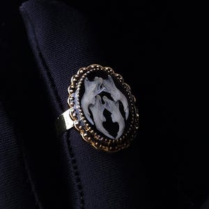 Könnte beinhalten: Goldfarbener Ring mit einem ovalen, schwarzen Mittelstück. Das Mittelstück zeigt eine Ansammlung weißer, organisch geformter Elemente. Der Ring hat einen dekorativen, gewellten Rand und ein schlichtes Band.