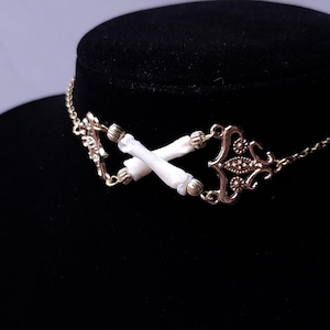 Puede incluir: Un collar gargantilla dorado con un dise&ntilde;o central de dos elementos blancos en forma de hueso cruzados. El collar tiene adornos de metal y una delicada cadena.