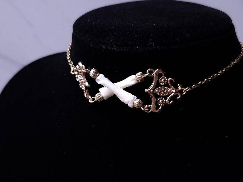 Puede incluir: Un collar gargantilla dorado con un dise&ntilde;o central de dos elementos blancos en forma de hueso cruzados. El collar tiene adornos de metal y una delicada cadena.