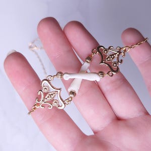 Puede incluir: Un collar dorado con un dise&ntilde;o delicado que presenta elementos de filigrana ornamentados y peque&ntilde;as piezas blancas con forma de hueso. El collar se sostiene en una mano, mostrando sus intrincados detalles y artesan&iacute;a. La cadena es fina y elegante.