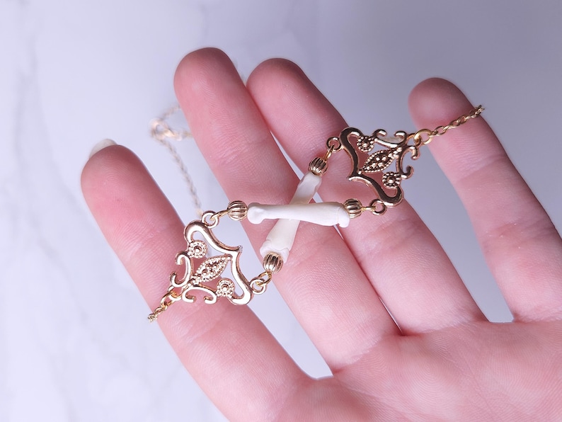 Puede incluir: Un collar dorado con un dise&ntilde;o delicado que presenta elementos de filigrana ornamentados y peque&ntilde;as piezas blancas con forma de hueso. El collar se sostiene en una mano, mostrando sus intrincados detalles y artesan&iacute;a. La cadena es fina y elegante.