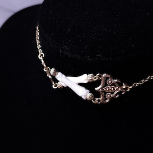 Puede incluir: Un collar dorado con una pieza central decorativa y detalles blancos. El collar presenta una cadena delicada y un colgante detallado con un dise&ntilde;o sim&eacute;trico. Los detalles blancos son cil&iacute;ndricos y a&ntilde;aden un toque &uacute;nico a la joya.