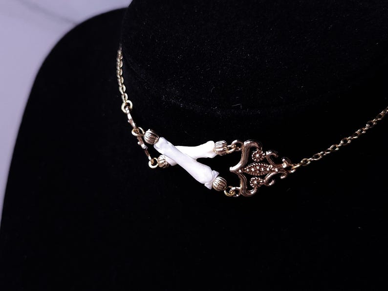 Puede incluir: Un collar dorado con una pieza central decorativa y detalles blancos. El collar presenta una cadena delicada y un colgante detallado con un dise&ntilde;o sim&eacute;trico. Los detalles blancos son cil&iacute;ndricos y a&ntilde;aden un toque &uacute;nico a la joya.