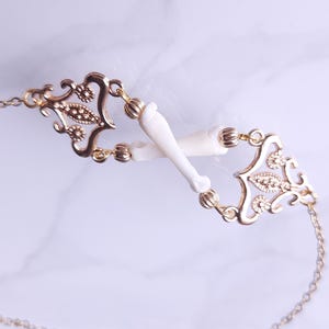 Puede incluir: Un collar dorado con una delicada cadena y detalles de filigrana ornamentados. El collar presenta dos elementos blancos en forma de hueso conectados por los eslabones dorados decorativos. El dise&ntilde;o es sim&eacute;trico y elegante.