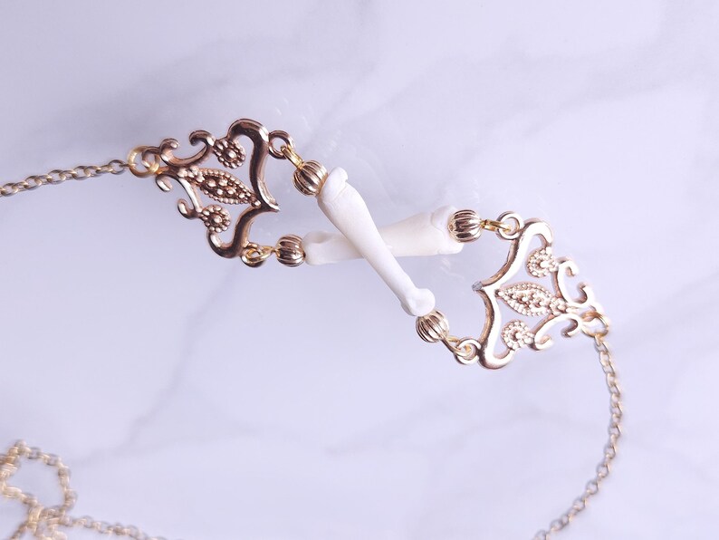 Puede incluir: Un collar dorado con una delicada cadena y detalles de filigrana ornamentados. El collar presenta dos elementos blancos en forma de hueso conectados por los eslabones dorados decorativos. El dise&ntilde;o es sim&eacute;trico y elegante.