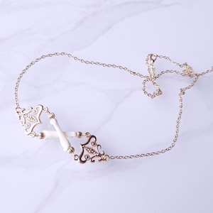 Puede incluir: Un collar dorado con una delicada cadena y colgantes ornamentados de estilo filigrana. El collar presenta detalles blancos en forma de hueso y se exhibe sobre una superficie de m&aacute;rmol. El dise&ntilde;o es sim&eacute;trico y elegante.