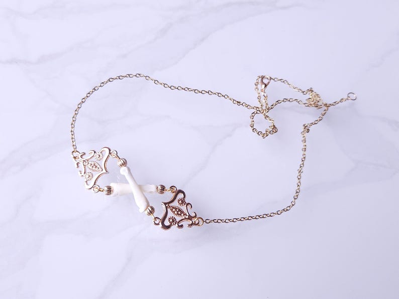 Puede incluir: Un collar dorado con una delicada cadena y colgantes ornamentados de estilo filigrana. El collar presenta detalles blancos en forma de hueso y se exhibe sobre una superficie de m&aacute;rmol. El dise&ntilde;o es sim&eacute;trico y elegante.
