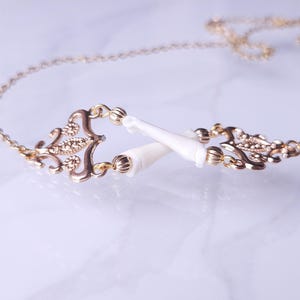 Puede incluir: Un collar dorado con un centro ornamental de estilo filigrana y dos elementos blancos puntiagudos. El collar tiene una cadena delicada. El fondo es claro.
