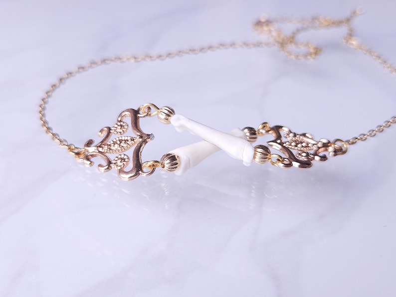 Puede incluir: Un collar dorado con un centro ornamental de estilo filigrana y dos elementos blancos puntiagudos. El collar tiene una cadena delicada. El fondo es claro.