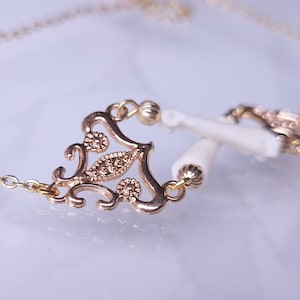 Puede incluir: Un collar dorado con un colgante de filigrana decorativo y una cadena delicada. El colgante presenta un dise&ntilde;o ornamentado con peque&ntilde;os acentos brillantes y est&aacute; conectado a dos elementos cil&iacute;ndricos blancos.