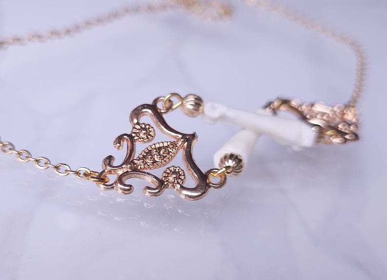 Puede incluir: Un collar dorado con un colgante de filigrana decorativo y una cadena delicada. El colgante presenta un dise&ntilde;o ornamentado con peque&ntilde;os acentos brillantes y est&aacute; conectado a dos elementos cil&iacute;ndricos blancos.