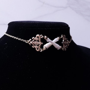 Puede incluir: Un collar gargantilla de color dorado con un dise&ntilde;o ornamentado. El collar presenta una cadena y elementos decorativos, incluido un dise&ntilde;o central con dos colgantes blancos en forma de hueso cruzados. El collar se exhibe en un maniqu&iacute; negro.