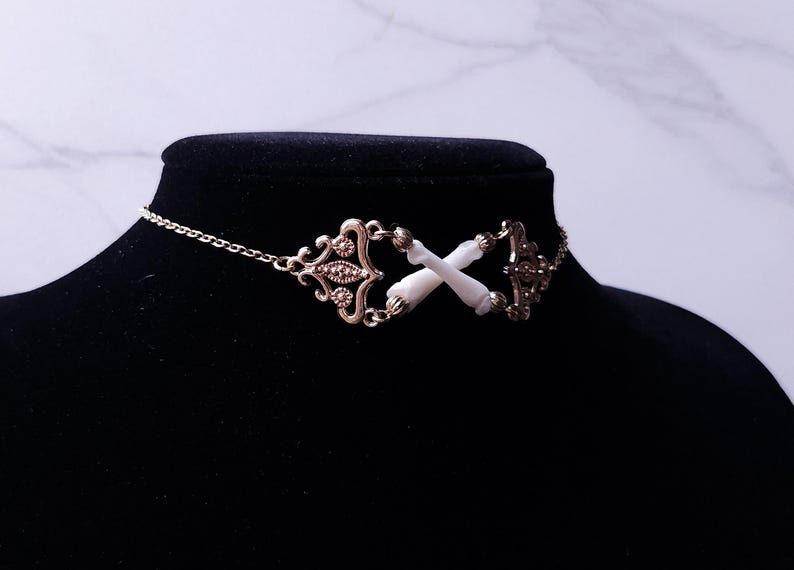 Puede incluir: Un collar gargantilla de color dorado con un dise&ntilde;o ornamentado. El collar presenta una cadena y elementos decorativos, incluido un dise&ntilde;o central con dos colgantes blancos en forma de hueso cruzados. El collar se exhibe en un maniqu&iacute; negro.