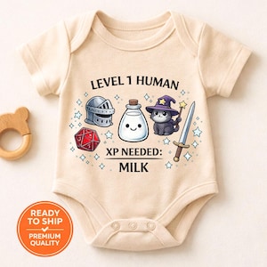 May include: A beige baby onesie with a playful graphic featuring a knight's helmet, a bottle of milk, a cat in a wizard hat, a sword, and a red die. Text reads "LEVEL 1 HUMAN" and "XP NEEDED: MILK".