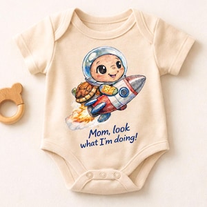 Può includere: Un body da bambino beige con un'illustrazione a fumetti di un bambino con un casco spaziale che cavalca un razzo. Il razzo è rosso, argento e blu, con fiamme. Il testo sul body recita: "Mamma, guarda cosa sto facendo!" A sinistra c'è un massaggiagengive in legno a forma di orso.