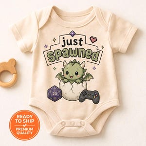 May include: Cream-colored baby onesie with the text "just spawned" above a cartoon dragon hatching from an egg. A purple die and a game controller are also featured. A wooden bear-shaped teether is on the left.