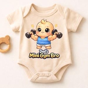 Puede incluir: Body de bebé beige con una ilustración de dibujos animados de un bebé levantando pesas. El bebé lleva una camiseta sin mangas azul y pantalones cortos blancos. El texto "Dad's Mini Gym Bro" está impreso debajo. A la izquierda, un mordedor de madera con forma de oso.