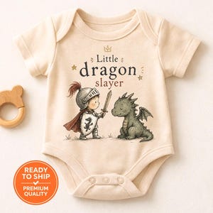 May include: A cream-coloured baby bodysuit featuring a cartoon knight and dragon illustration, with the text "Little dragon slayer". A wooden bear-shaped teether is to the left. The bodysuit is made of soft material.