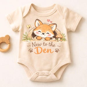 May include: Cream-colored baby onesie with a cartoon fox illustration. The fox is nestled in a den, with the text "New to the Den" below. A wooden bear-shaped teether is to the left.