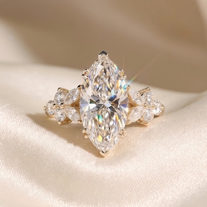 Op de afbeelding: Een gouden ring met een grote, marquise-geslepen diamant als middelpunt, geflankeerd door kleinere, bloemblad-vormige stenen. De band en zetting van de ring zijn goud, wat een luxueuze en elegante uitstraling creëert. De ring wordt gepresenteerd op een zacht, crèmekleurig oppervlak.