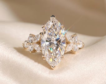 Puede incluir: Un anillo de oro con un gran diamante de talla marquesa en el centro, flanqueado por piedras más pequeñas en forma de pétalo. La banda y el engaste del anillo son de oro, creando una apariencia lujosa y elegante. El anillo se muestra sobre una superficie crema.