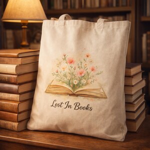 Peut inclure: Un sac fourre-tout beige avec un motif floral et le texte "Lost In Books". Le motif présente un livre ouvert avec des fleurs roses et blanches. Le sac a deux poignées et est posé sur une surface en bois.