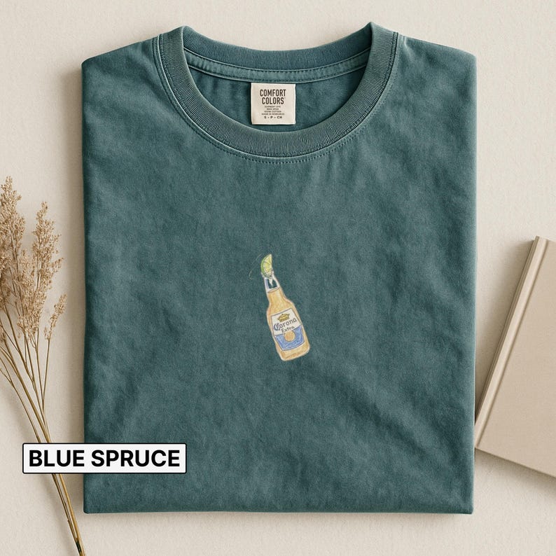 Puede incluir: Camiseta azul abeto con un peque&ntilde;o bordado de una botella de cerveza Corona Extra con una rodaja de lima. La camiseta tiene cuello redondo y la etiqueta de la marca Comfort Colors. El texto "BLUE SPRUCE" est&aacute; en un rect&aacute;ngulo blanco en la parte inferior.