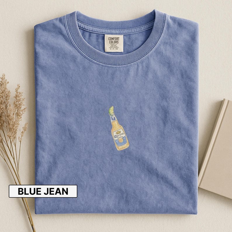 Puede incluir: Camiseta azul vaquero con un peque&ntilde;o bordado de una botella de cerveza Corona con una rodaja de lima. La camiseta est&aacute; doblada y tiene una etiqueta en el cuello. El texto "BLUE JEAN" est&aacute; en un rect&aacute;ngulo blanco en la parte inferior.