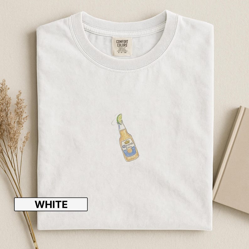 Puede incluir: Camiseta blanca con cuello redondo. La camiseta presenta una botella de cerveza Corona bordada con una rodaja de lima. La palabra "WHITE" est&aacute; en un rect&aacute;ngulo blanco en la parte inferior de la imagen. Hecho por Comfort Colors.