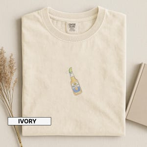 Puede incluir: Camiseta de color marfil con cuello redondo. La camiseta presenta una peque&ntilde;a y colorida ilustraci&oacute;n de una botella de cerveza Corona con una rodaja de lima. La camiseta tiene una etiqueta que dice "Comfort Colors". La palabra "IVORY" est&aacute; impresa en un rect&aacute;ngulo blanco.