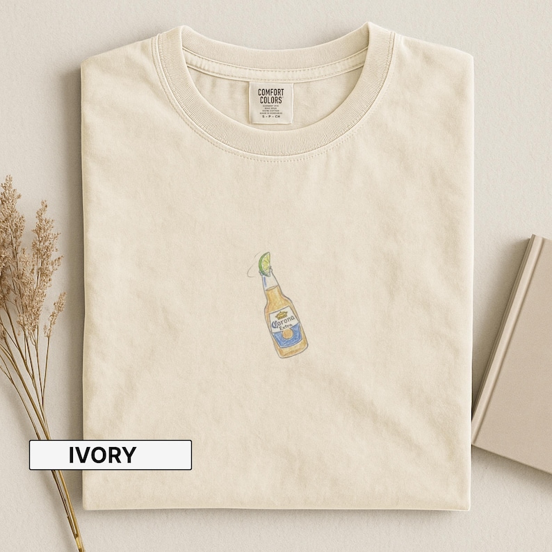 Puede incluir: Camiseta de color marfil con cuello redondo. La camiseta presenta una peque&ntilde;a y colorida ilustraci&oacute;n de una botella de cerveza Corona con una rodaja de lima. La camiseta tiene una etiqueta que dice "Comfort Colors". La palabra "IVORY" est&aacute; impresa en un rect&aacute;ngulo blanco.