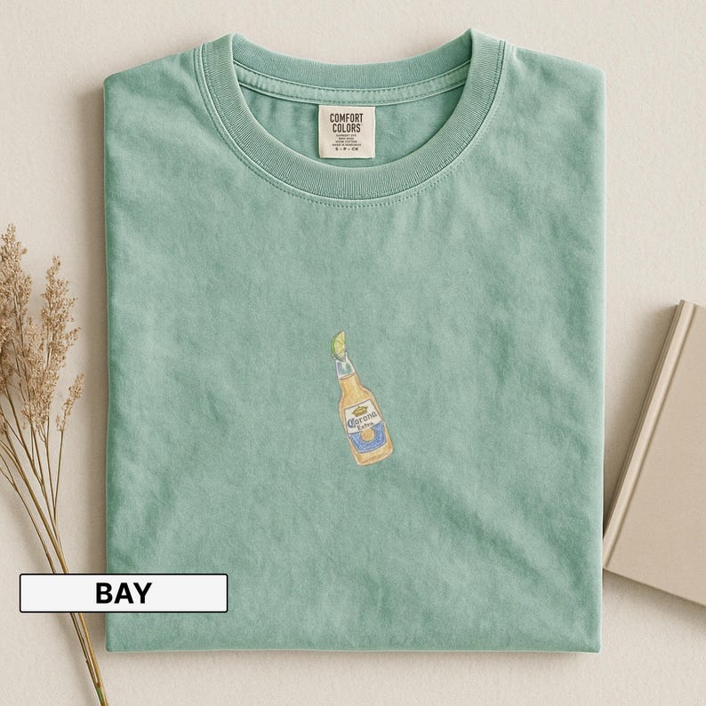 Puede incluir: Camiseta verde claro con cuello redondo y mangas cortas. La camiseta presenta una ilustraci&oacute;n bordada de una botella de cerveza Corona Extra con una rodaja de lima. La etiqueta dice "Comfort Colors".