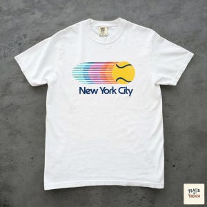 May include: A white short-sleeve t-shirt featuring a graphic of a yellow tennis ball with a multi-coloured motion trail in shades of blue, purple, pink, and orange. Below the graphic, the text New York City is printed in a dark blue font.