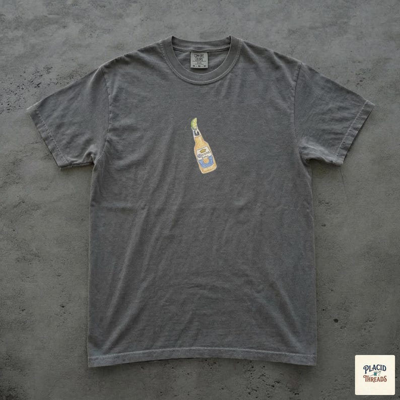 Puede incluir: Una camiseta gris con un peque&ntilde;o gr&aacute;fico de una botella de cerveza. La botella de cerveza es amarilla con detalles azules y blancos. La camiseta tiene cuello redondo y mangas cortas. La etiqueta de la marca es visible en la parte superior de la camiseta.