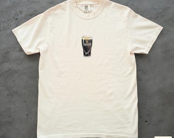 Op de afbeelding: Crèmekleurig T-shirt met een kleine afbeelding van een pint Guinness. Het bierglas is donker met een schuimende kop. Het shirt heeft korte mouwen en een ronde hals. Het Plaid Threads-logo staat rechtsonder.