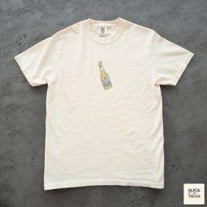 Puede incluir: Camiseta de color crema con un peque&ntilde;o gr&aacute;fico de una botella de cerveza con una rodaja de lima. La botella tiene una etiqueta azul y amarilla. La camiseta est&aacute; hecha de un material suave y c&oacute;modo.