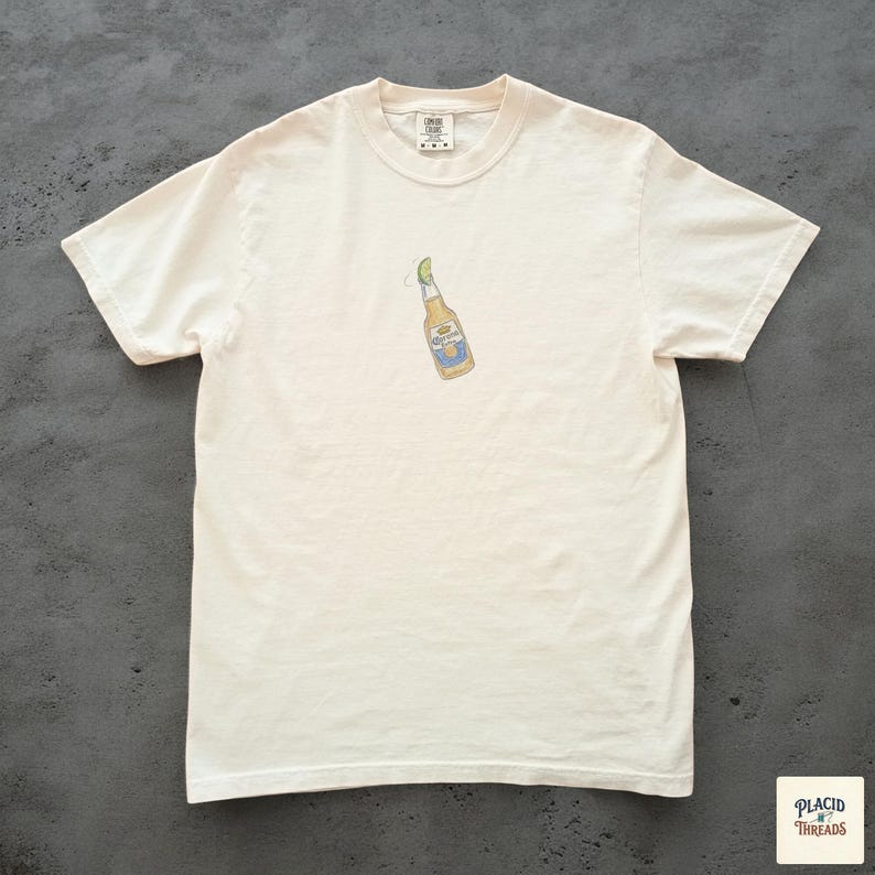 Puede incluir: Camiseta de color crema con un peque&ntilde;o gr&aacute;fico de una botella de cerveza con una rodaja de lima. La botella tiene una etiqueta azul y amarilla. La camiseta est&aacute; hecha de un material suave y c&oacute;modo.