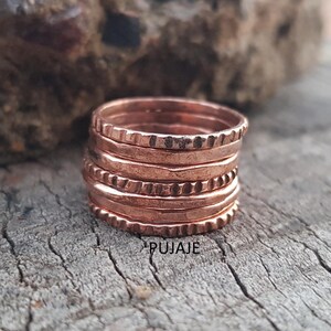 Peut inclure: Une pile de bagues de couleur cuivre avec divers motifs texturés, dont des motifs martelés et perlés. Les bagues sont empilées sur une surface en bois vieilli. Le mot "PUJAJE" est imprimé sur le bois.