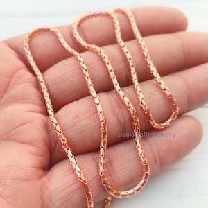Puede incluir: Una cadena de collar en tono oro rosa con un diseño de eslabones entrelazados texturizados y un patrón repetitivo en forma de escamas. La superficie metálica tiene un brillo metálico y el texto poojacraftedjewelry es visible cerca del centro.